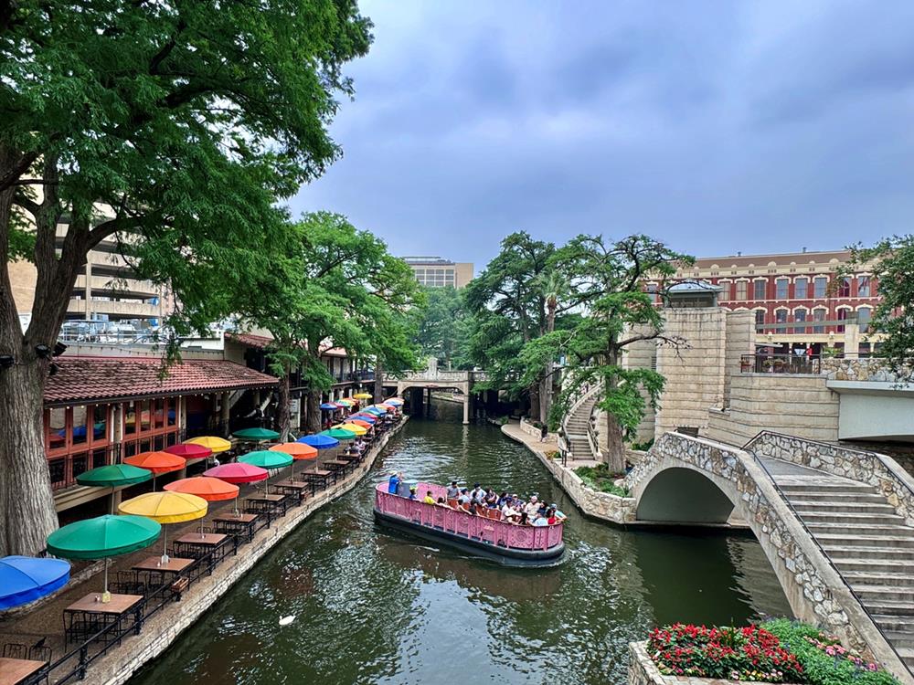 A scenic view of the San Antonio River Walk featuring a riverboat cruise passing under historic bridges, lined with colorful umbrellas and lush greenery